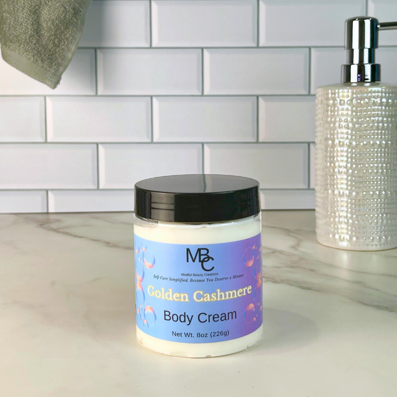A jar of MBC Golden Cashmere Body Cream with a black lid and purple label, set on a marble countertop in front of a white subway tile backsplash, with a soap dispenser in the background.