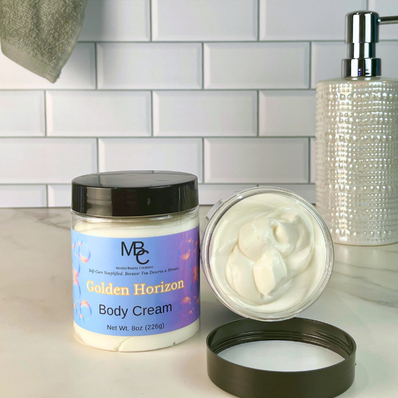 A jar of MBC Golden Horizon Body Cream is shown on a bathroom counter with the lid off, revealing the thick white cream inside. A matching textured soap dispenser and white subway tile backsplash are visible in the background.