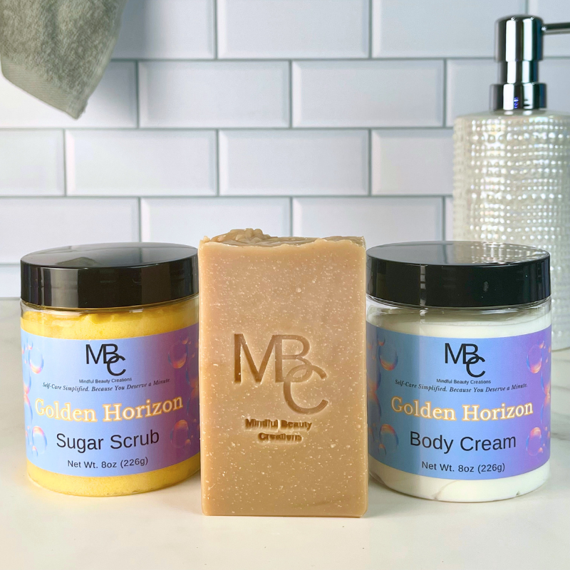 Three MBC Beauty Care products on a white countertop against a white subway tile backsplash: a orange jar of Golden Horizon Sugar Scrub, a tan bar of soap in the center, and a white jar of Golden Horizon Body Cream. A soap dispenser and hanging towel are partially visible in the background.