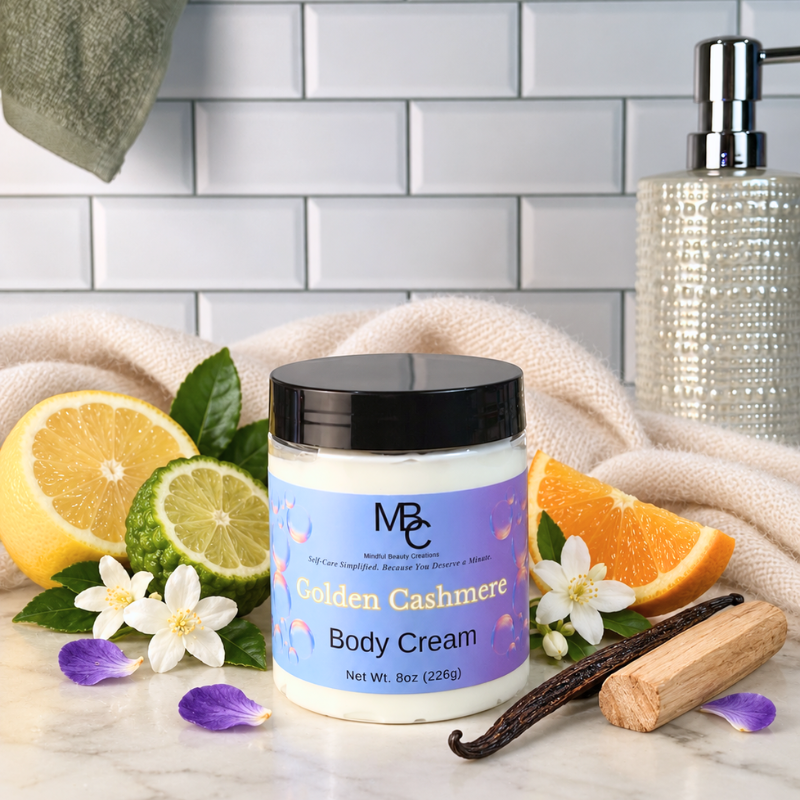 A jar of MBC Golden Cashmere Body Cream is displayed on a marble surface with bath-themed props including a soap dispenser, a towel, citrus slices, vanilla bean, and flowers against a white subway tile background.