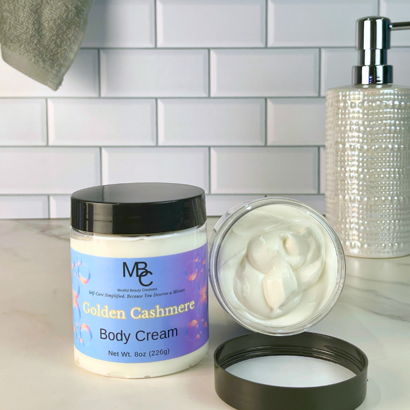 A jar of MBC Golden Cashmere Body Cream shown with its lid off, revealing the thick white cream inside. The product is set on a white marble countertop with a white subway tile backsplash, a textured silver soap dispenser, and a gray towel in the background.