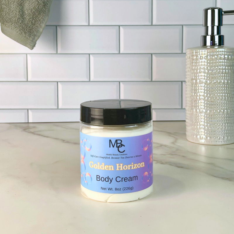 A jar of MBC Golden Horizon Body Cream sits on a white marble countertop in front of a white subway tile wall. A textured white soap dispenser is visible on the right, and a gray towel hangs on the left.