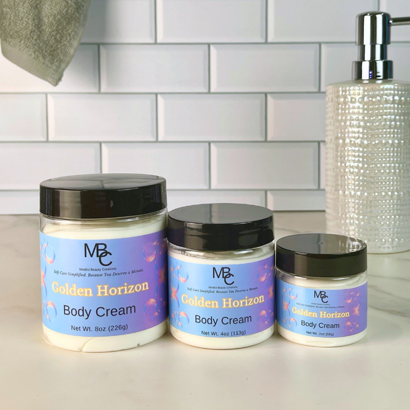Three jars of Mindful Beauty Creations Golden Horizon Body Cream in different sizes, lined up on a white countertop with a white subway tile backsplash behind them.