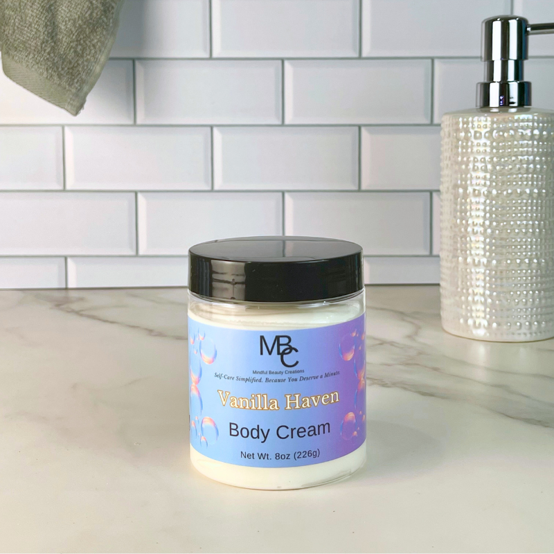 A jar of MBC Body Cream labeled 'Vanilla Haven' sits on a marble countertop in front of a white subway tile backsplash. A textured white soap dispenser is visible on the right, and a gray towel hangs in the upper left.