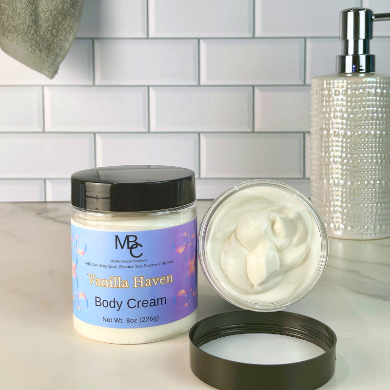 A jar of MBC Vanilla Haven Body Cream shown open on a countertop, with the cream visible in the clear container and the lid placed beside it. A white subway-tile backsplash and a textured silver pump bottle are in the background.
