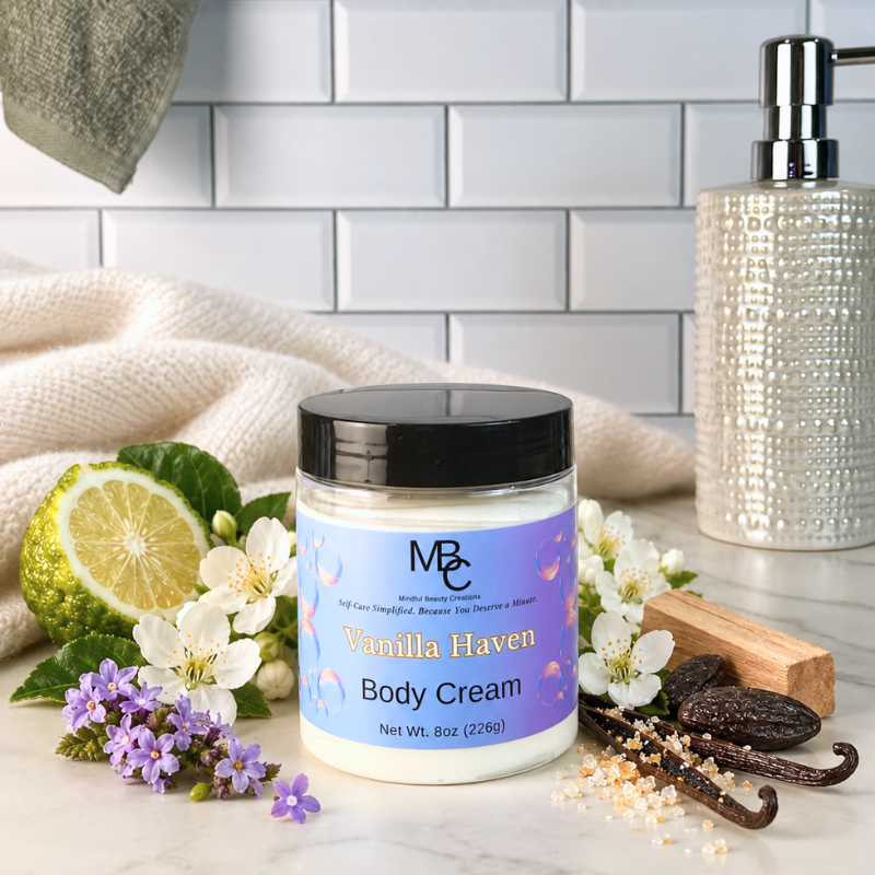 A jar of MBC Vanilla Haven Body Cream is styled on a marble surface with a lime, white blossoms, vanilla beans, and bath accessories against a white subway tile background.