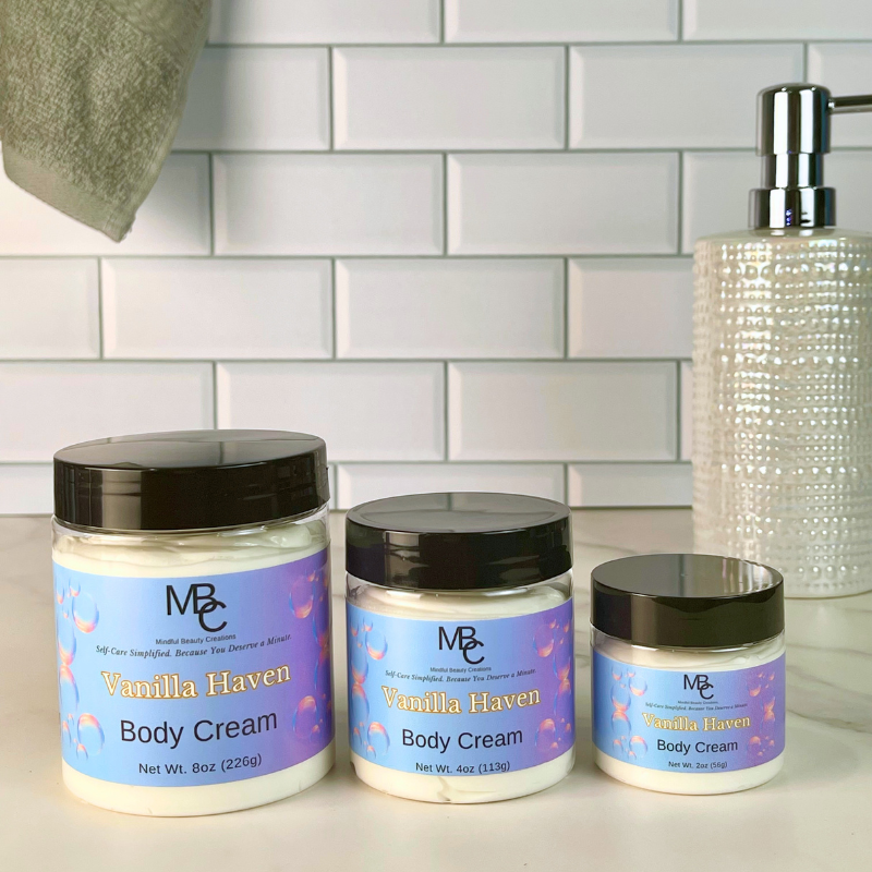 Three jars of Mindful Beauty Creations 'Vanilla Haven' body cream in different sizes arranged on a white countertop against a white subway tile backsplash. A textured clear pump bottle and a folded gray towel are visible in the background.