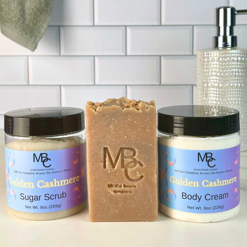 Three self-care products from Mindful Beauty Creations arranged on a white surface against a white subway tile backsplash: a jar of Golden Cashmere Sugar Scrub, a bar of Golden Cashmere Body Butter, and a jar of Golden Cashmere Body Cream. A textured soap dispenser is partially visible behind the products.