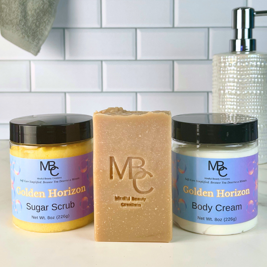 Three MBC Beauty Care products on a white countertop against a white subway tile backsplash: a orange jar of Golden Horizon Sugar Scrub, a tan bar of soap in the center, and a white jar of Golden Horizon Body Cream. A soap dispenser and hanging towel are partially visible in the background.