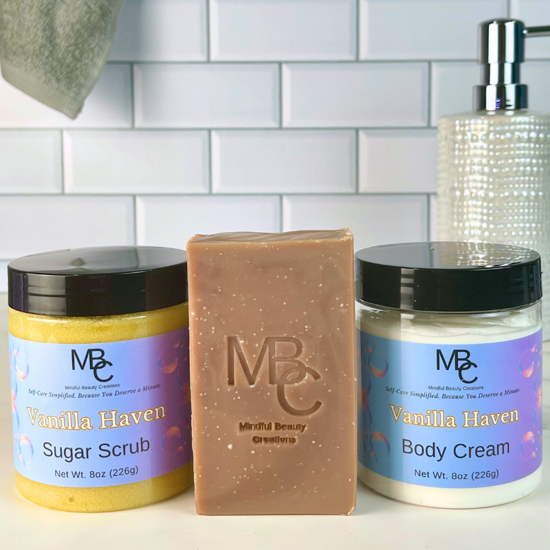 Three MBC beauty products from the Vanilla Haven line are arranged on a white surface against a white subway tile backsplash: a brown jar of Sugar Scrub, a brown bar of Body Cream, and a white jar of Body Cream. A textured soap dispenser is partially visible behind the products.