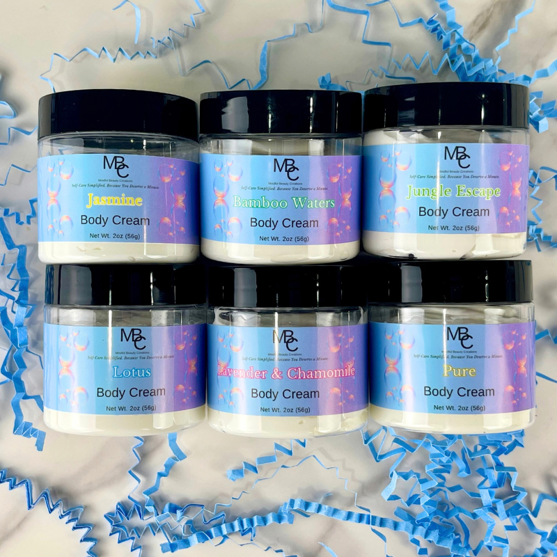 Six 2oz jars of Mindful Beauty Creations Body Cream arranged in two rows on a white marble surface with blue crinkle paper. The jars have black lids and colorful labels featuring a purple-to-blue gradient with bubbles and text for different scents: Jasmine, Bamboo Waters, Jungle Escape, Lotus, Lavender & Chamomile, and Pure.