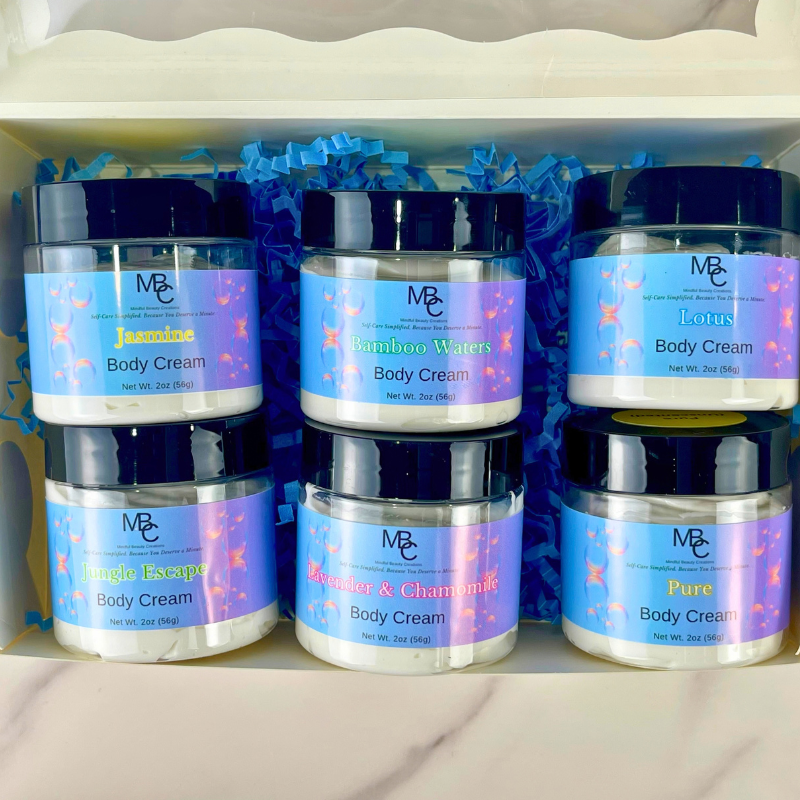 Six 2oz jars of Mindful Beauty Creations Body Cream arranged in a gift box with blue crinkle paper. The labels feature designs with blue, purple, and pink bubble patterns, and the visible scents include Jasmine, Lotus, Jungle Escape, Bamboo Waters, Lavender & Chamomile, and Pure.