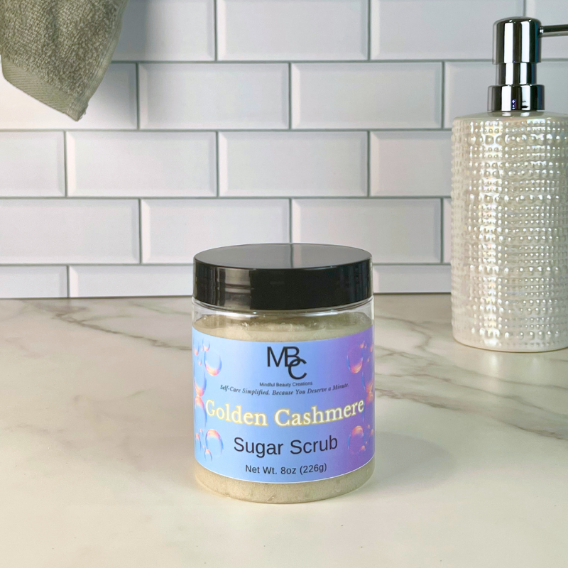 A jar of MBC 'Golden Cashmere' sugar scrub sits on a white marble countertop in a bathroom setting, with a white subway tile backsplash behind it and a textured soap dispenser to the right.