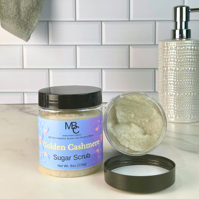 A jar of MBC Golden Cashmere Sugar Scrub is shown on a bathroom counter next to an open jar displaying the creamy scrub texture, with a tiled wall and soap dispenser in the background.