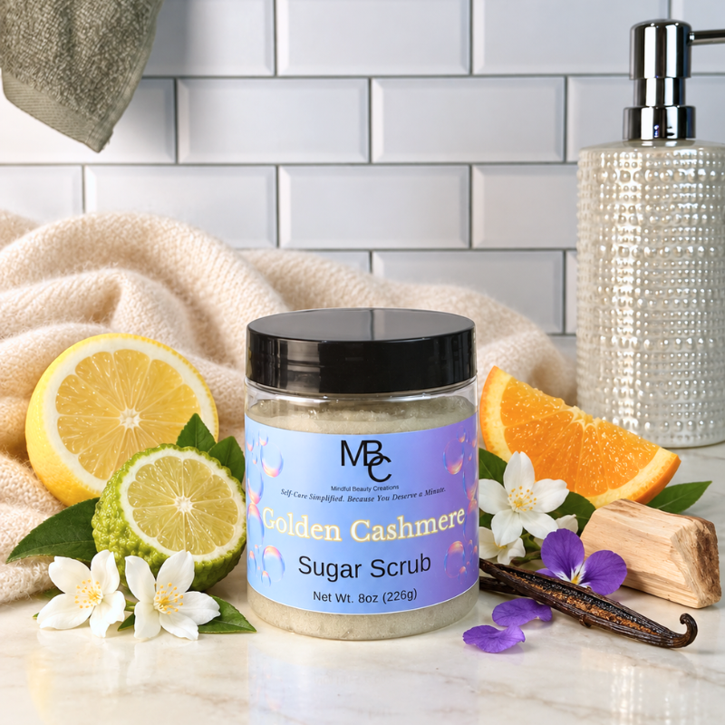 A jar of MBC Golden Cashmere Sugar Scrub sits on a marble countertop surrounded by citrus slices, white flowers, a vanilla bean, and a soap dispenser against a white subway tile background.