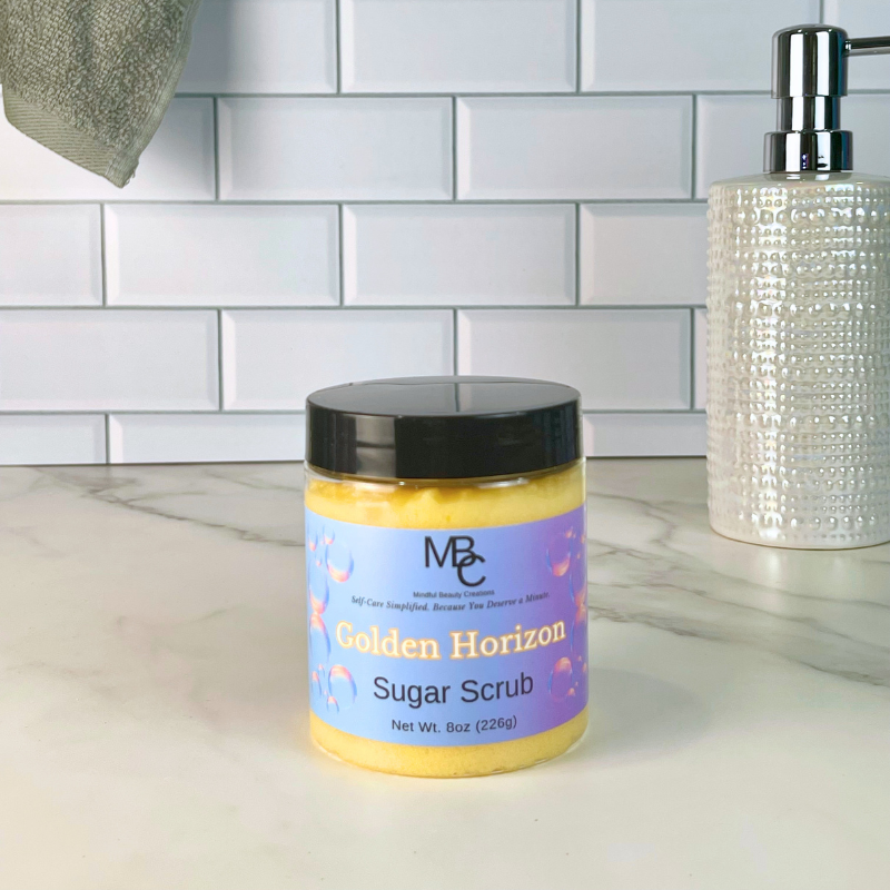 A jar of Mindful Beauty Creations Golden Horizon Sugar Scrub with a black lid and purple label, sitting on a white marble countertop in front of a white subway tile wall.