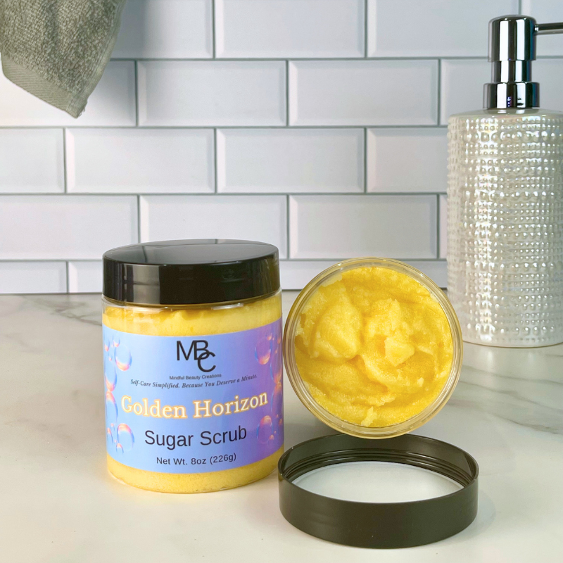 A jar of MBC Golden Horizon Sugar Scrub with its lid off, showing the orange scrub inside, set on a marble countertop with a tiled backsplash background.
