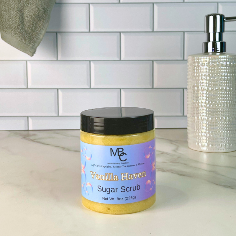 A jar of MBC Vanilla Haven Sugar Scrub sits on a marble countertop in front of a white subway tile backsplash. A textured white soap dispenser is visible on the right, and a gray towel hangs on the left.
