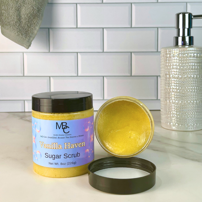 A jar of MBC Vanilla Haven Sugar Scrub shown on a bathroom counter with the lid off, revealing the brown scrub inside, alongside a soap dispenser and tiled wall background.