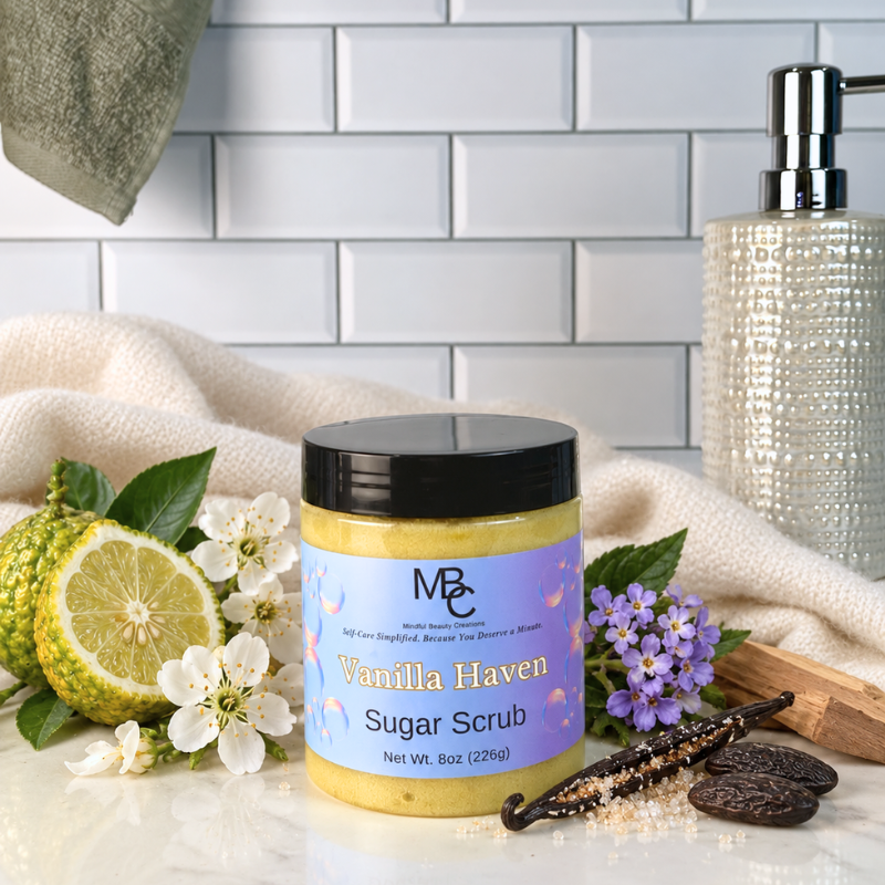 A jar of MBC Beauty product 'Vanilla Haven' sugar scrub, styled with a sliced lime, white flowers, a vanilla bean, and sugar crystals on a marble countertop with a white subway tile backsplash in the background.