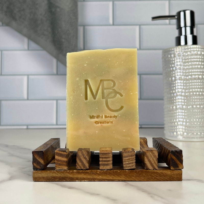 Bar of Coastal Bloom soap with 'MBC' branding on a wooden soap dish in a bathroom setting.
