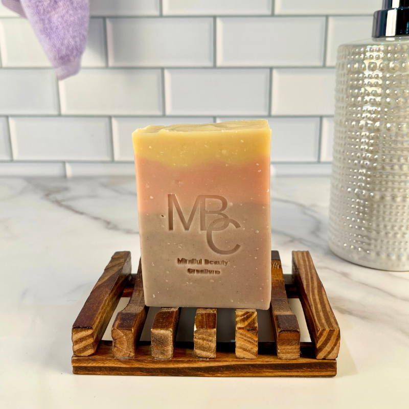 Yellow, pink, and purple Bar of soap with 'MBC' branding on a wooden soap dish against a tiled bathroom wall.