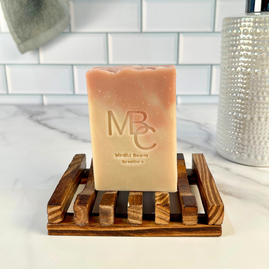 Pink and tan Bar of soap with 'MBC' branding on a wooden soap dish against a bathroom background.