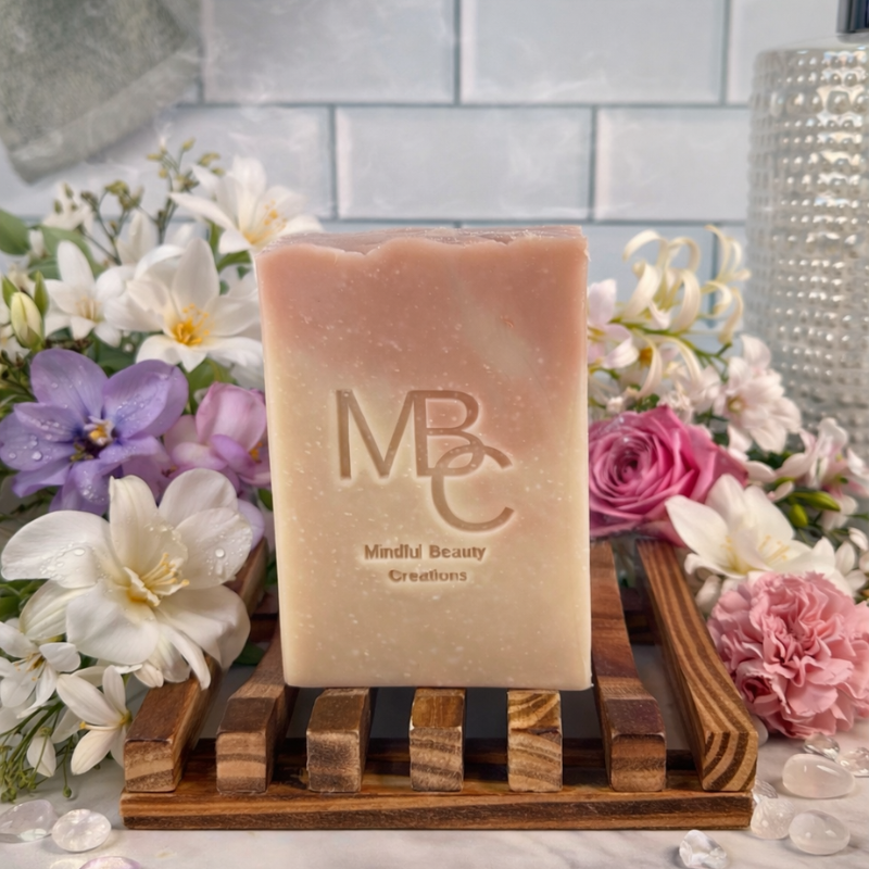 Pink and tan Bar of soap with 'MBC' branding on a wooden stand surrounded by flowers.