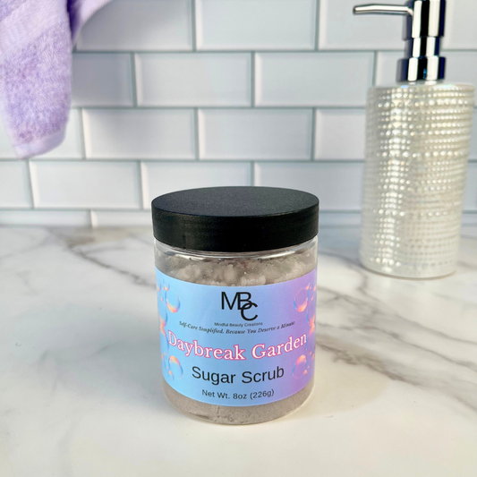 Jar of Daybreak Garden Sugar Scrub on a bathroom counter with soap dispenser and towel in the background.