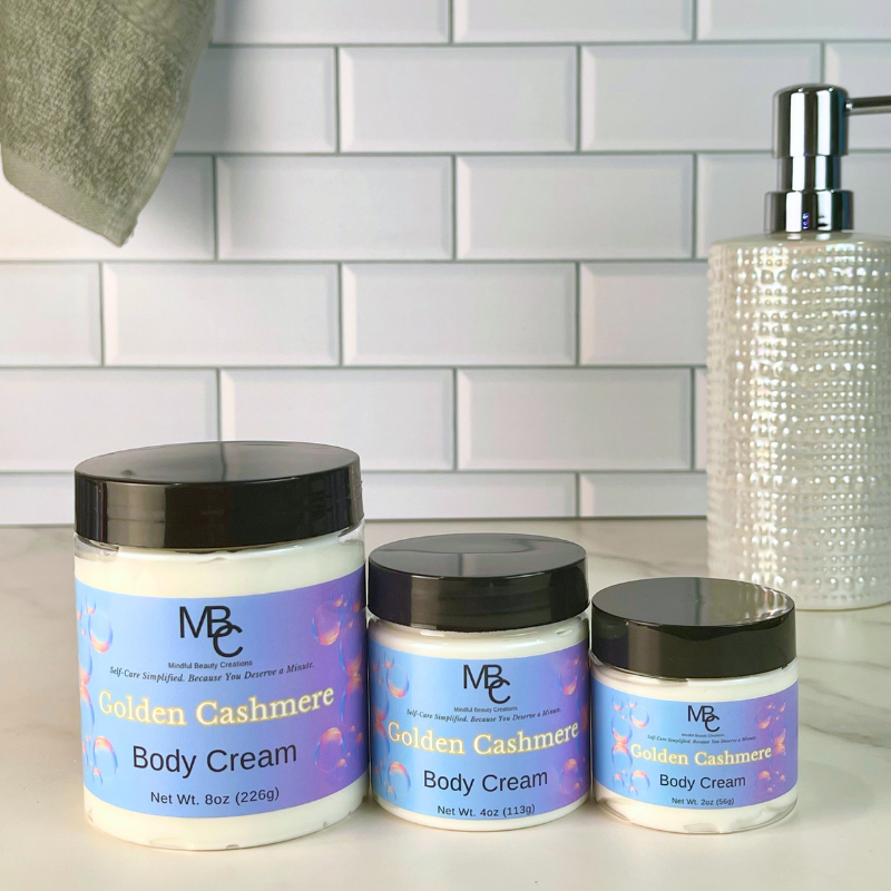 Three jars of MBC Self Care 'Golden Cashmere' body cream in different sizes, lined up on a white countertop against a white subway tile backsplash. A textured clear pump bottle is visible in the background on the right.