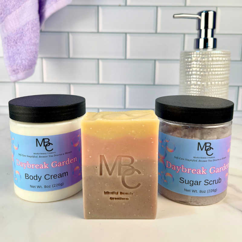 Three beauty products including a jar of body cream, a bar of soap, and a jar of sugar scrub on a tiled bathroom counter. All are named Daybreak Garden.