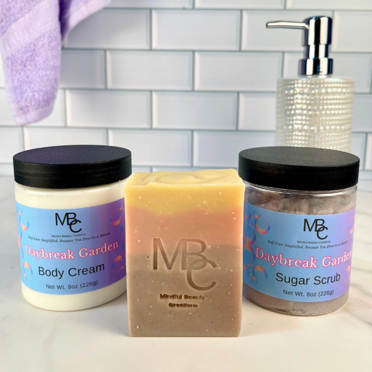 Three beauty products including a jar of body cream, a bar of soap, and a jar of sugar scrub on a tiled bathroom counter. All are named Daybreak Garden.