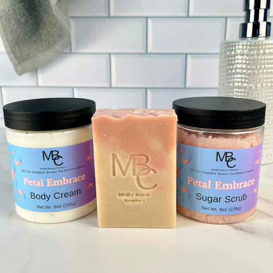 Three beauty products including a jar of body cream, a bar of soap, and a jar of sugar scrub on a bathroom counter. All are named Petal Embrace.