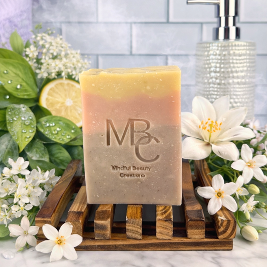 Yellow, pink, and purple Bar of soap with 'MBC' branding on a wooden soap dish, surrounded by flowers and greenery.