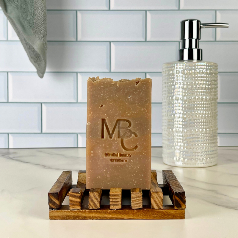A rectangular bar of brown soap standing on a wooden soap dish, with a clear textured pump bottle behind it against a white subway tile backsplash.