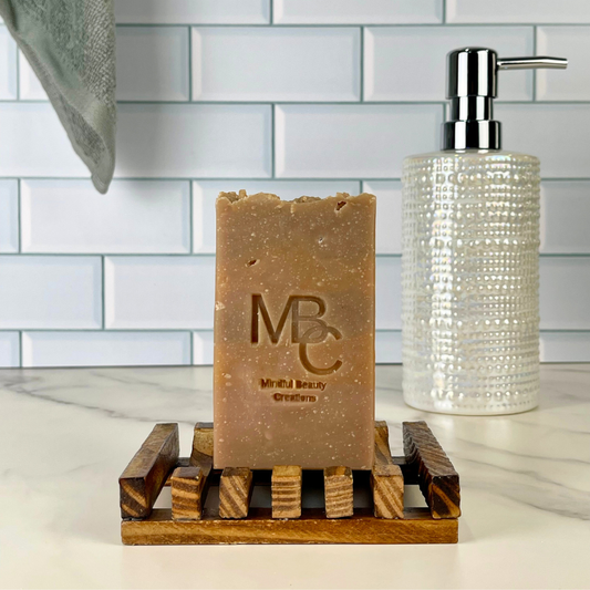 A rectangular bar of brown soap standing on a wooden soap dish, with a clear textured pump bottle behind it against a white subway tile backsplash.