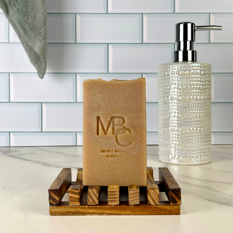 A rectangular tan soap bar with embossed 'MBC' branding sits on a small wooden soap dish, with a textured clear pump bottle and a white subway-tile backsplash in the background.
