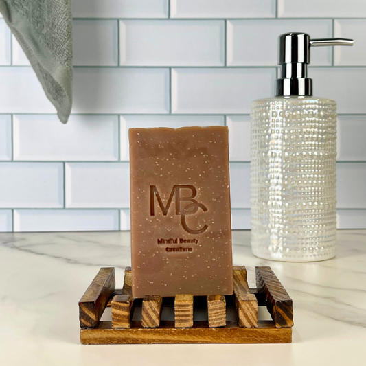 A rectangular bar of brown soap with white speckles and embossed 'MBC' branding sits on a wooden soap dish. A clear textured pump bottle is behind it against a white subway tile backsplash.